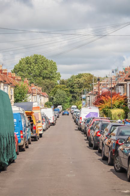 A residential street in Finsbury Park with rows of parked cars lining both sides of the road, including compact cars, vans, and utility vehicles, some with covers or protective wrapping, indicative of a home relocation or moving process. The street features terraced houses with brick facades, small front gardens, and trees with green and red foliage, under an overcast sky with gray clouds. Power lines run above the street. In the background, there are larger trees and houses, suggesting a suburban neighbourhood. The scene portrays an environment suitable for furniture transport and packing during a house removal, with vehicles parked close to the properties’ front entrances, supporting local moving services such as those offered by Man with Van Finsbury Park.