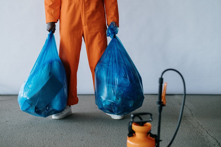 Close-up of a person dressed in orange work trousers and white sneakers, holding two large blue plastic bags filled with waste. The individual is standing on a grey carpeted floor inside a property, w