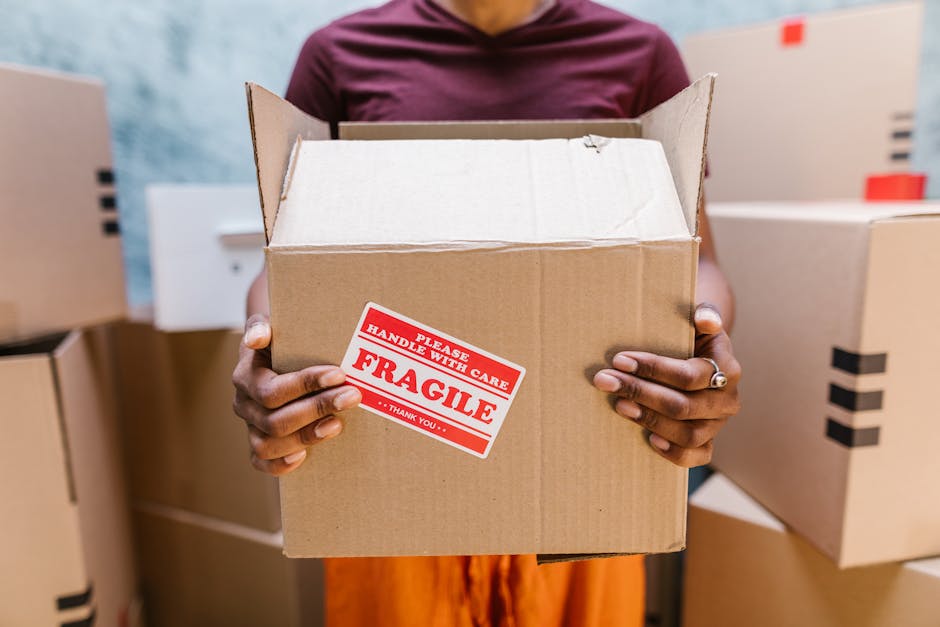 A person wearing a white t-shirt and blue jeans is kneeling inside a room, filling out a label on a cardboard moving box with a red marker. The box, sealed with white packing tape, has the word 'Spare