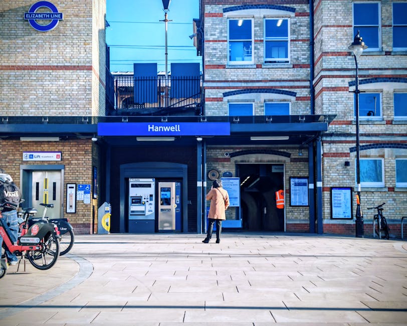 A person dressed in a beige coat and dark pants stands outside the entrance to Hanwell station, part of the Elizabeth Line, with a small group of bicycles parked on the pavement nearby. The station's brick building features large windows with blue frames, a black canopy with the station name 'Hanwell' displayed in white letters, and a sign indicating lift access. The scene is illuminated by natural daylight, and adjacent to the station entrance, there are digital information boards, a black lamppost, and a black bike rack. To the left, there is an ATM machine embedded into the brick wall. The setting suggests a location suitable for home relocation or furniture transport, with ongoing assistance from services like Man with Van Finsbury Park, supporting efficient packing and moving logistics in an urban environment.