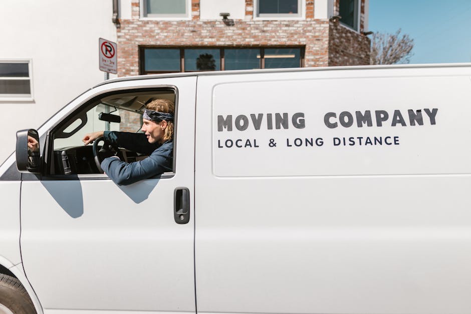 A white moving van parked outside a residential building with a brick facade and large windows, featuring the logo and text 'MOVING COMPANY LOCAL & LONG DISTANCE' on the side. Inside the cab, a person with long, light brown hair tied back, wearing a dark blue jacket and a black headband, is sitting in the driver's seat, preparing to start the vehicle. The vehicle is positioned on the pavement near the building's entrance, with a no-parking sign visible in the background. The scene captures the loading or transport phase of a home relocation, indicating furniture transport and moving logistics related to house removals, as promoted by Man with Van Finsbury Park.
