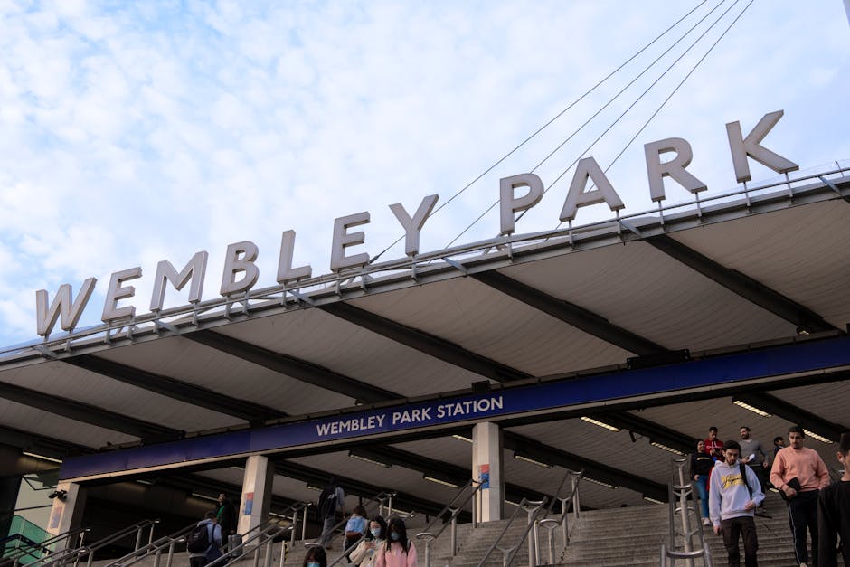A person dressed in a beige coat and dark pants stands outside the entrance to Hanwell station, part of the Elizabeth Line, with a small group of bicycles parked on the pavement nearby. The station's brick building features large windows with blue frames, a black canopy with the station name 'Hanwell' displayed in white letters, and a sign indicating lift access. The scene is illuminated by natural daylight, and adjacent to the station entrance, there are digital information boards, a black lamppost, and a black bike rack. To the left, there is an ATM machine embedded into the brick wall. The setting suggests a location suitable for home relocation or furniture transport, with ongoing assistance from services like Man with Van Finsbury Park, supporting efficient packing and moving logistics in an urban environment.