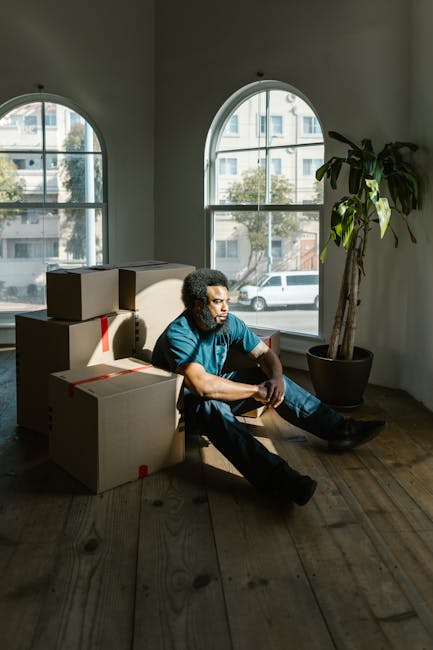 A man with curly hair and a beard, wearing a blue t-shirt and dark trousers, sits on a wooden floor inside a room with white walls and large arched windows that let in natural light. He is surrounded by several cardboard boxes of various sizes, some sealed with red packing tape, indicating they are packed for a house move or relocation. The boxes are stacked around him, several positioned near a large potted plant with broad green leaves. Outside the windows, a city street with parked cars and buildings is visible. The room appears to be in the process of being packed or emptied, reflecting home packing and moving activities, with the man taking a moment during a furniture transport or home relocation process. The scene conveys the preparation phase of a professional removals service, such as that offered by [COMPANY_NAME], as part of a house removal or furniture transport operation.