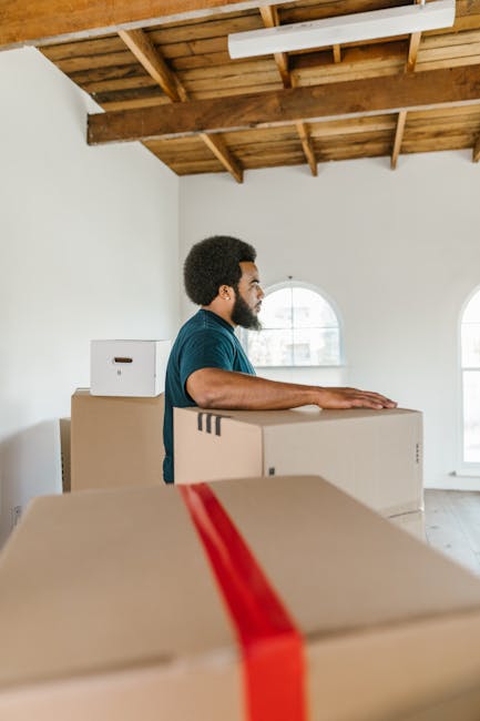 A man with curly hair and a beard, wearing a blue t-shirt and dark trousers, sits on a wooden floor inside a room with white walls and large arched windows that let in natural light. He is surrounded by several cardboard boxes of various sizes, some sealed with red packing tape, indicating they are packed for a house move or relocation. The boxes are stacked around him, several positioned near a large potted plant with broad green leaves. Outside the windows, a city street with parked cars and buildings is visible. The room appears to be in the process of being packed or emptied, reflecting home packing and moving activities, with the man taking a moment during a furniture transport or home relocation process. The scene conveys the preparation phase of a professional removals service, such as that offered by [COMPANY_NAME], as part of a house removal or furniture transport operation.